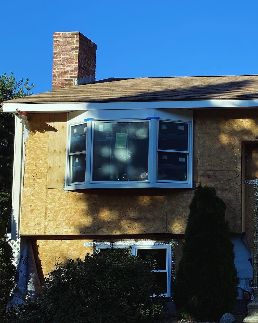 Close-up of a home exterior during renovation, featuring a newly installed bay window set into exposed plywood sheathing, with blue tape around the window trim and sunlight casting shadows across the unfinished siding.