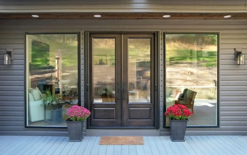 Exterior entry area with double glass doors and adjacent large window, set in gray siding with recessed porch lighting, potted flowers, and a welcoming deck surface.