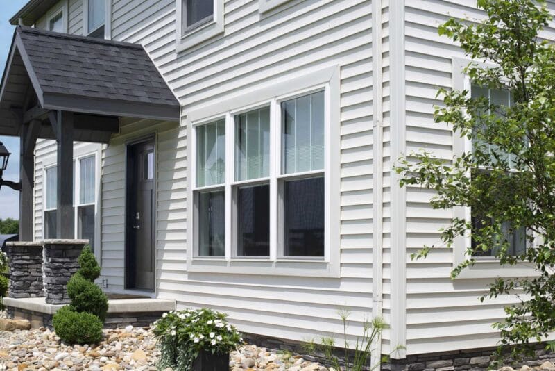 Exterior of a white vinyl-sided home featuring a black front door under a small covered entry, double-hung windows with white trim, stone porch columns, and landscaped shrubs and stone ground cover along the foundation.