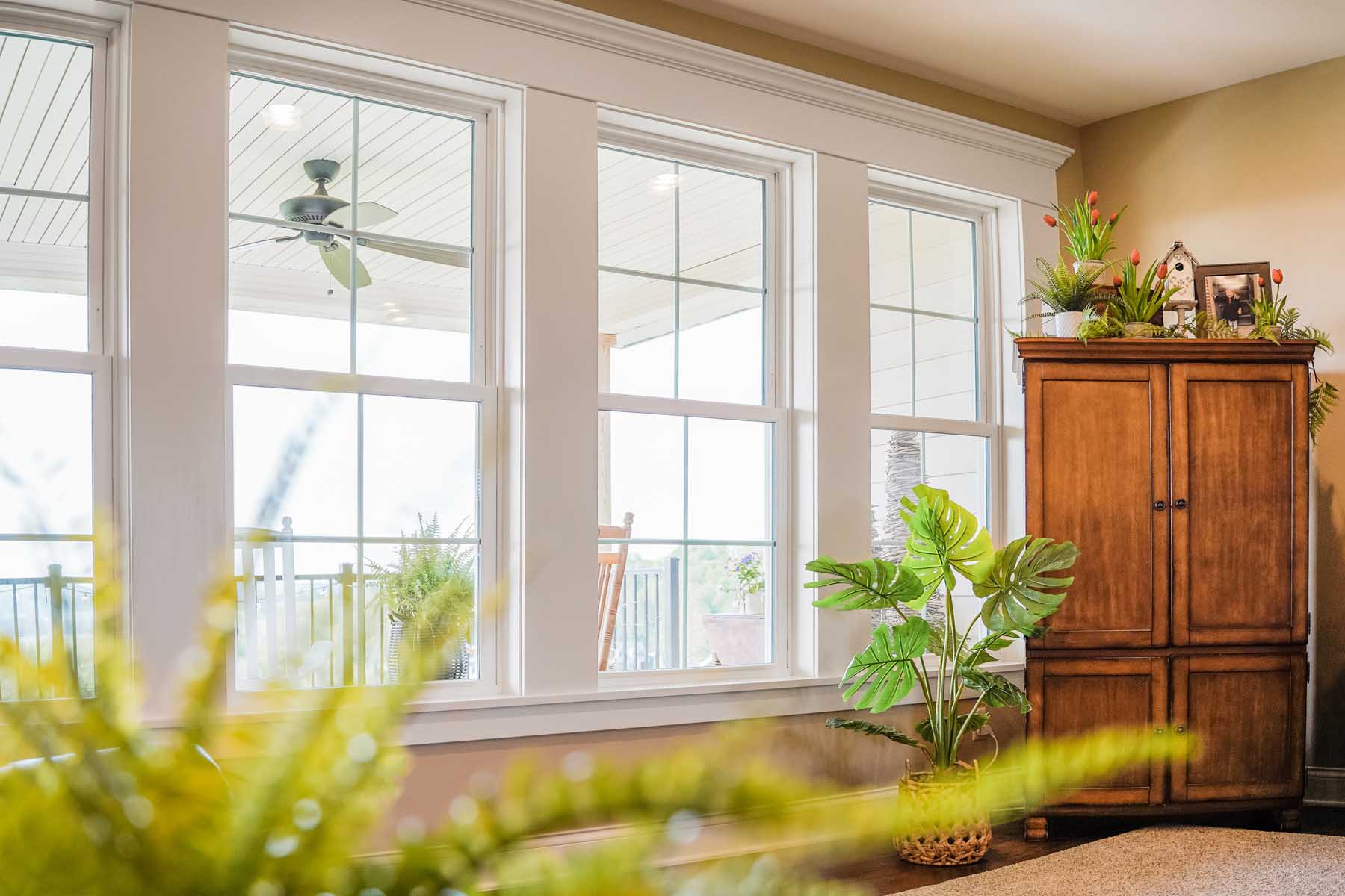 Interior view of three large white-trimmed windows bringing in natural light, with a porch ceiling fan visible outside and houseplants and a wooden cabinet arranged along the wall.