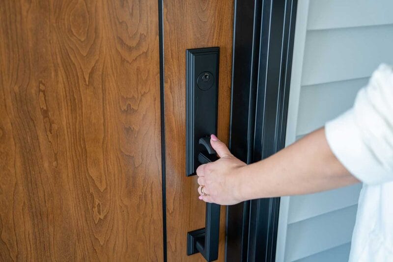 Close-up of a hand gripping a matte black entry door handle and lockset on a wood-grain front door with a dark trim surround.