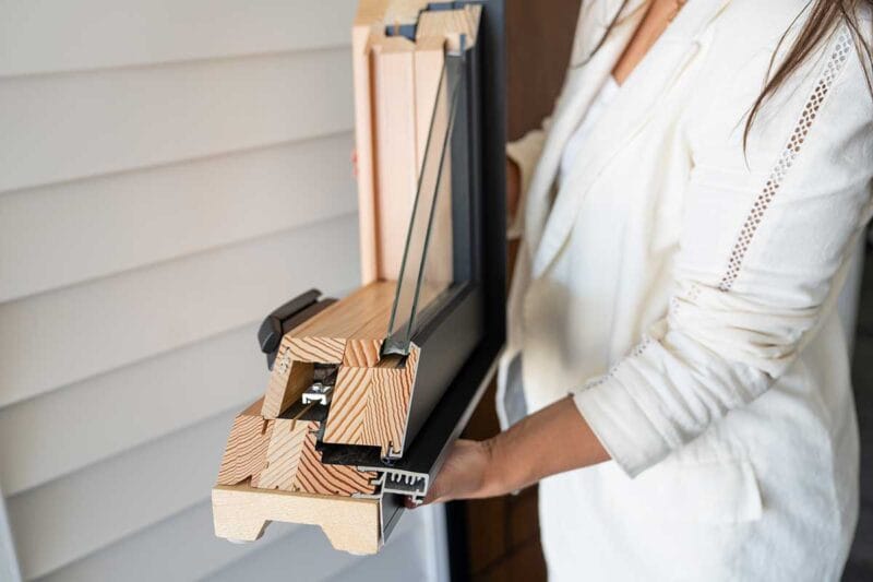 Cross-section view of a wood-framed window held by a person, showing layered wood joinery, insulated glass panes, weather seals, and an aluminum exterior cladding profile.