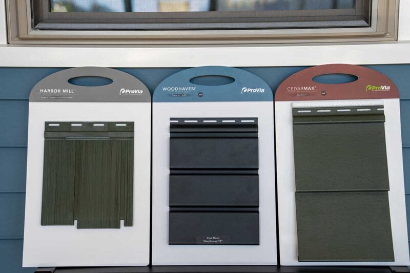 Three exterior siding sample boards displayed side by side beneath a window, showing green vertical panels, black horizontal panels, and dark green horizontal panels.