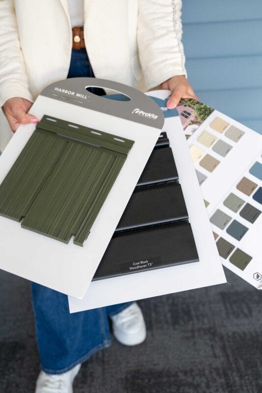 Person holding exterior siding and color sample boards, including green and black panel samples, with a swatch booklet showing multiple neutral color options.