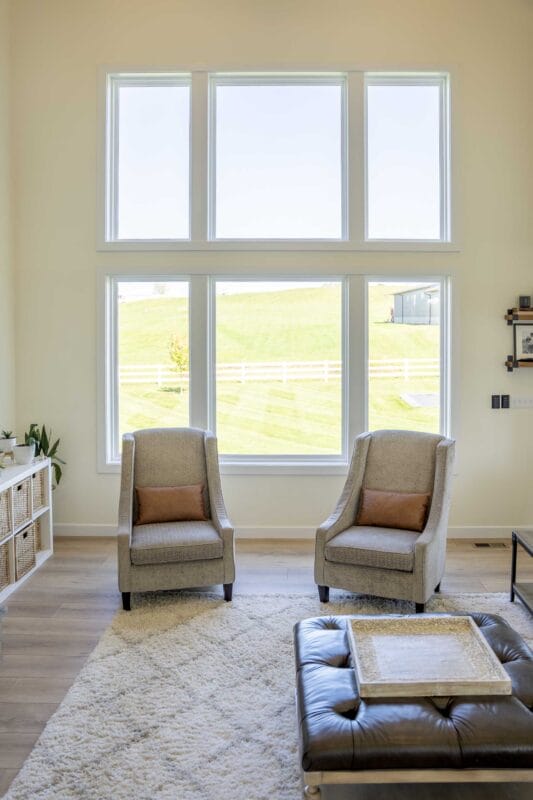 Bright living room with tall stacked windows overlooking a grassy landscape, two upholstered armchairs on a light area rug, and a leather ottoman coffee table in the foreground.