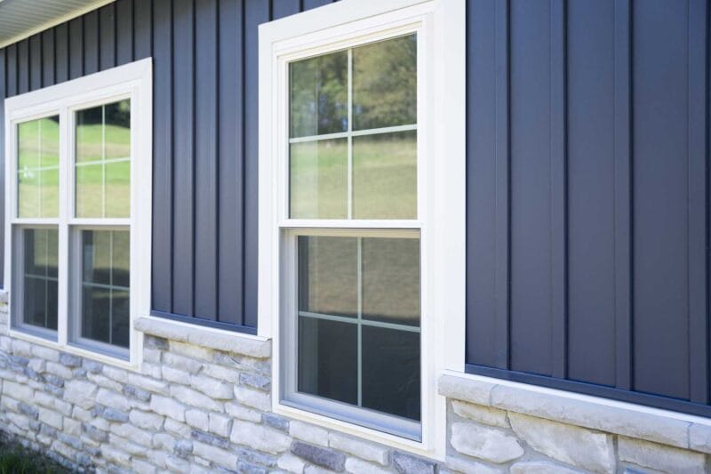 Exterior view of a home with dark vertical siding, white-trimmed double-hung windows, and a stone veneer foundation reflecting a grassy landscape.