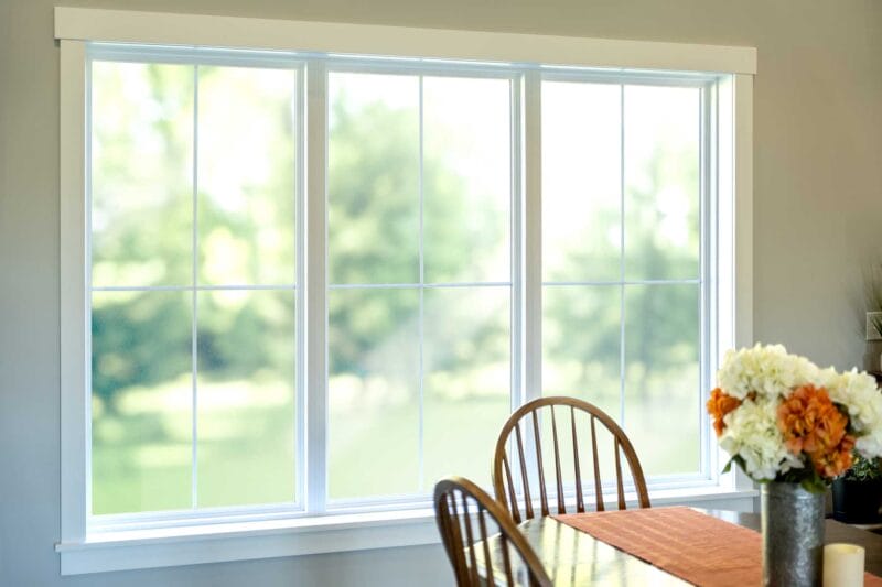 Wide three-panel window with white trim and grid panes, letting in soft natural light and a blurred green outdoor view beside a wooden dining table with flowers.