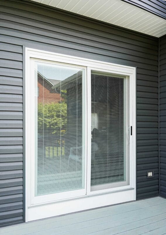 White sliding glass patio door with built-in blinds, set in gray horizontal siding beneath a covered porch ceiling, opening onto a light gray deck.
