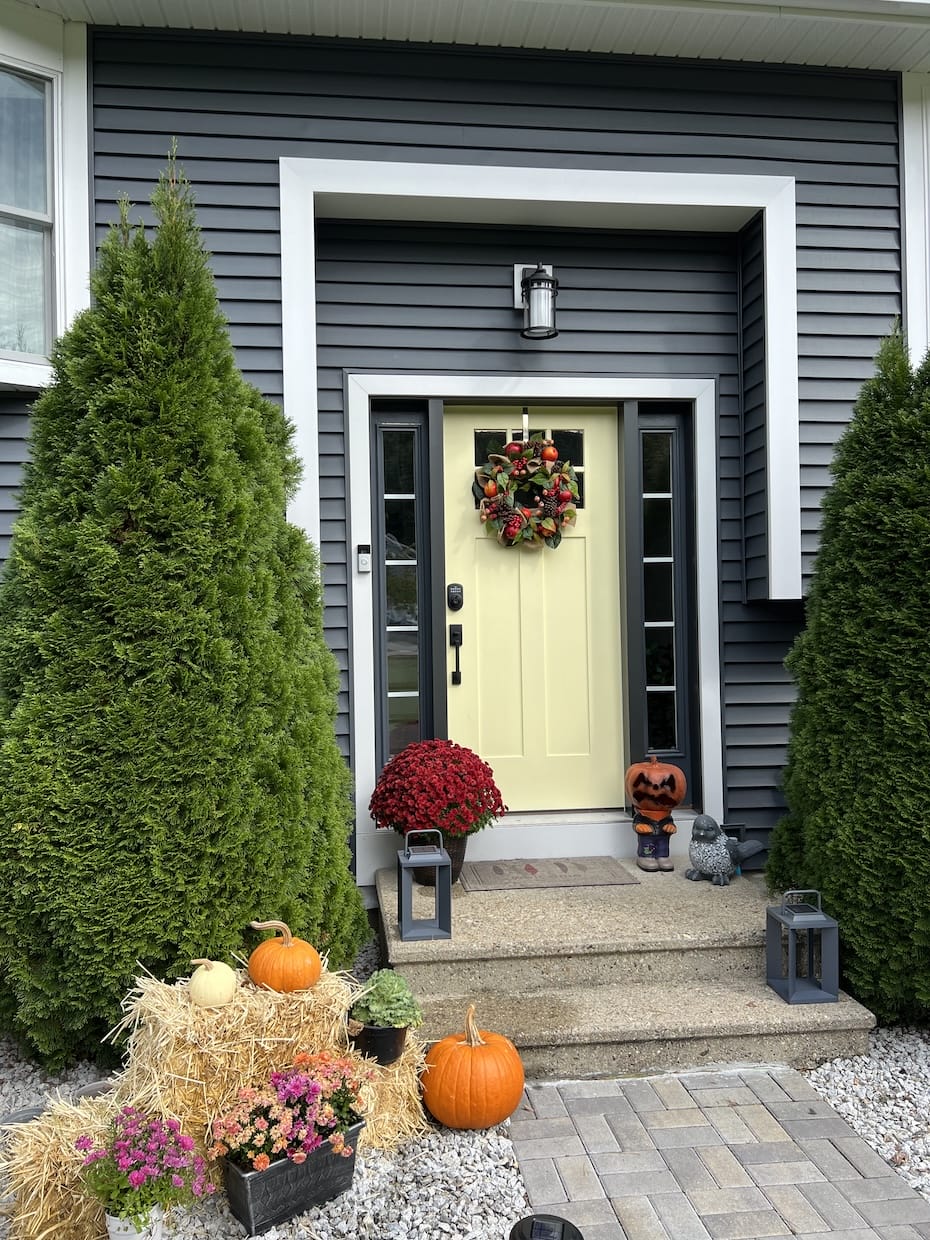 A front entryway with dark gray siding and white trim, featuring a pale yellow door decorated with a fall wreath, pumpkins, mums, hay bales, lanterns, and evergreen shrubs framing the steps.