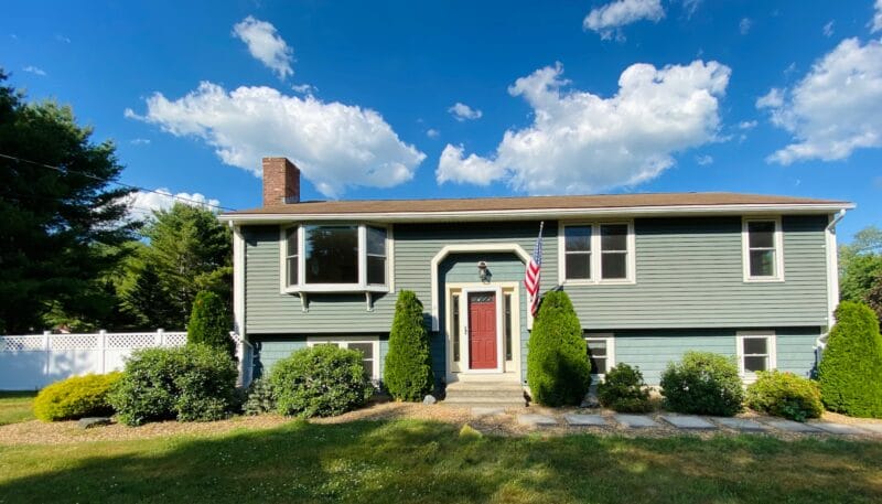 A well-maintained split-level home with green siding, landscaped front yard, and an American flag, set beneath a bright blue sky with mature trees surrounding the property.