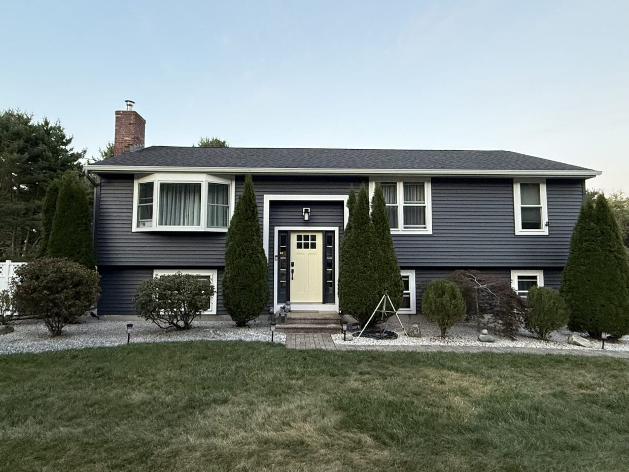 A split-level home with dark gray siding, white trim, and a centered pale yellow front door, framed by evergreen shrubs, gravel landscaping, and a neatly maintained lawn.