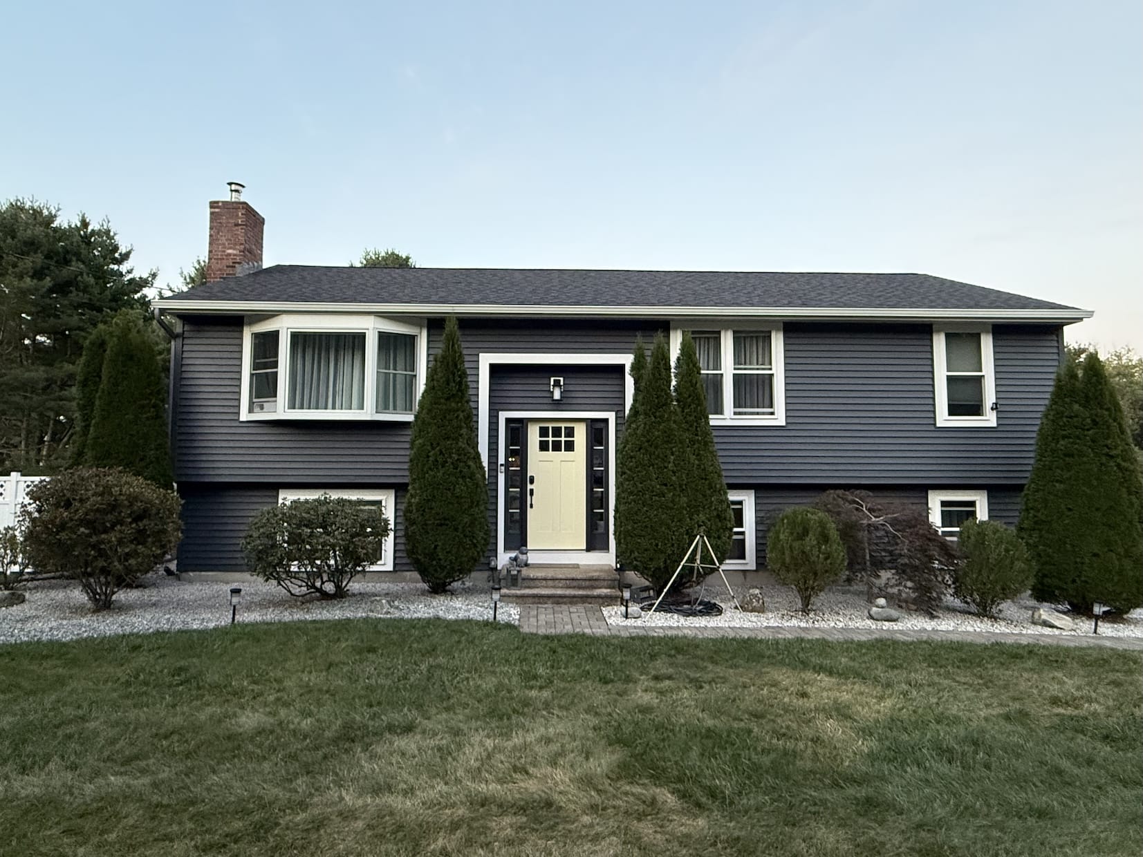 A split-level home with dark gray siding, white trim, and a centered pale yellow front door, framed by evergreen shrubs, gravel landscaping, and a neatly maintained lawn.