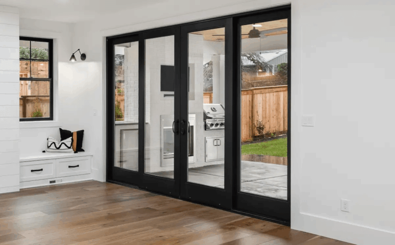 Interior living space featuring black-framed multi-panel sliding glass doors opening to a covered patio with an outdoor kitchen, hardwood floors, and a built-in window bench.