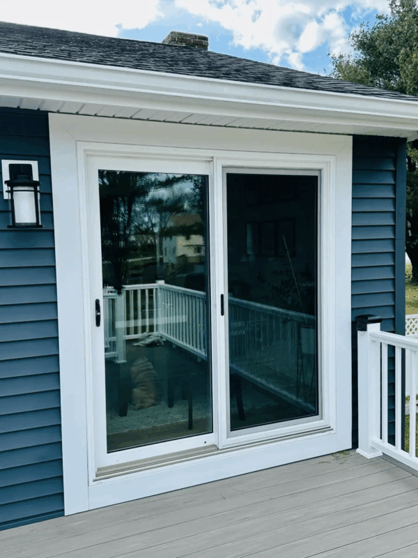 White sliding glass patio door installed in blue siding with wide white trim, opening onto a deck with white railing beneath a roof overhang.