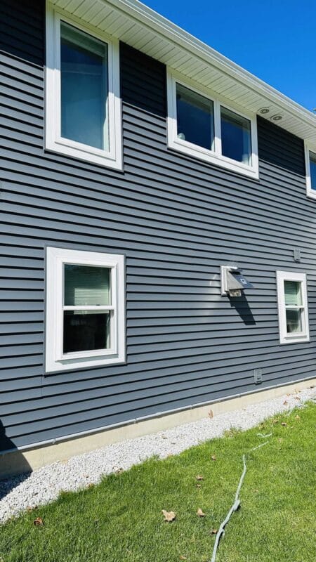 Side view of a two-story home with dark blue horizontal siding, white-trimmed windows on both levels, white soffits, gravel along the foundation, and green lawn under a clear blue sky.
