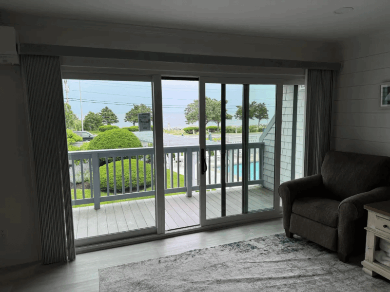 Interior living room with a wide sliding glass door opening to a balcony, featuring vertical blinds, coastal views, and natural light filling the space.
