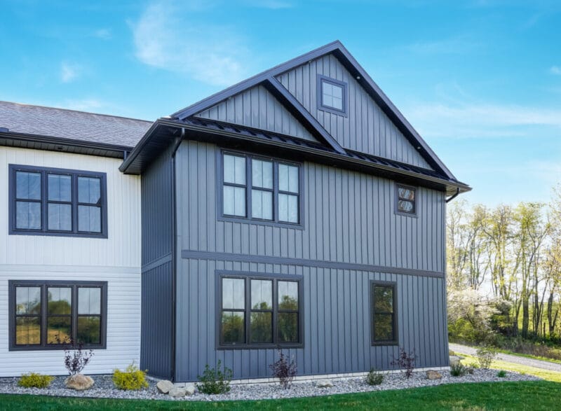 Rear exterior of a modern home with blue-gray board-and-batten siding, black-trimmed windows, a gable roof with metal accents, and stone landscaping under a clear sky.