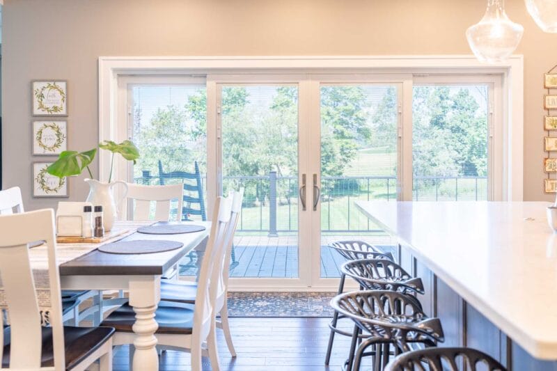 Bright dining area with a large white sliding glass door opening to a deck, featuring a farmhouse-style table, white chairs, and natural light throughout the space.