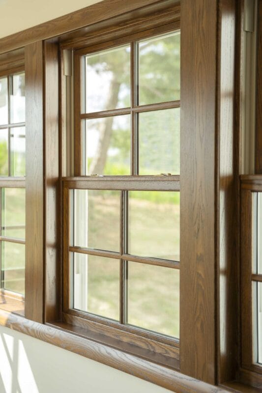 Close-up of an interior wood-framed window with a warm stained finish, divided panes, and sunlight highlighting the trim and sill with a soft outdoor view beyond.