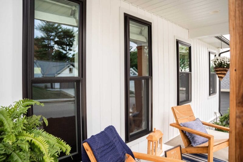 Front porch with white vertical siding and a row of black-framed windows, furnished with wooden chairs, blue cushions, potted plants, and hanging flowers overlooking a quiet neighborhood.