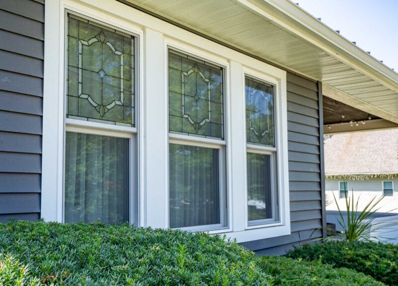 Exterior view of three white-trimmed double-hung windows with decorative glass inserts on a gray-sided home, framed by shrubs and a covered porch roof.
