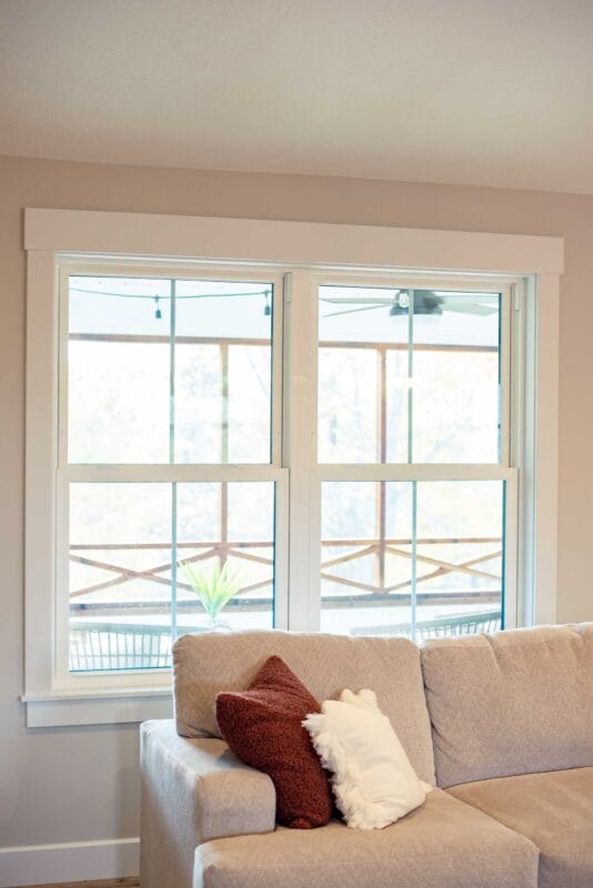 Living room window with white trim behind a beige sectional sofa, showing a covered porch outside with wooden railing, string lights, and a ceiling fan visible through the glass.