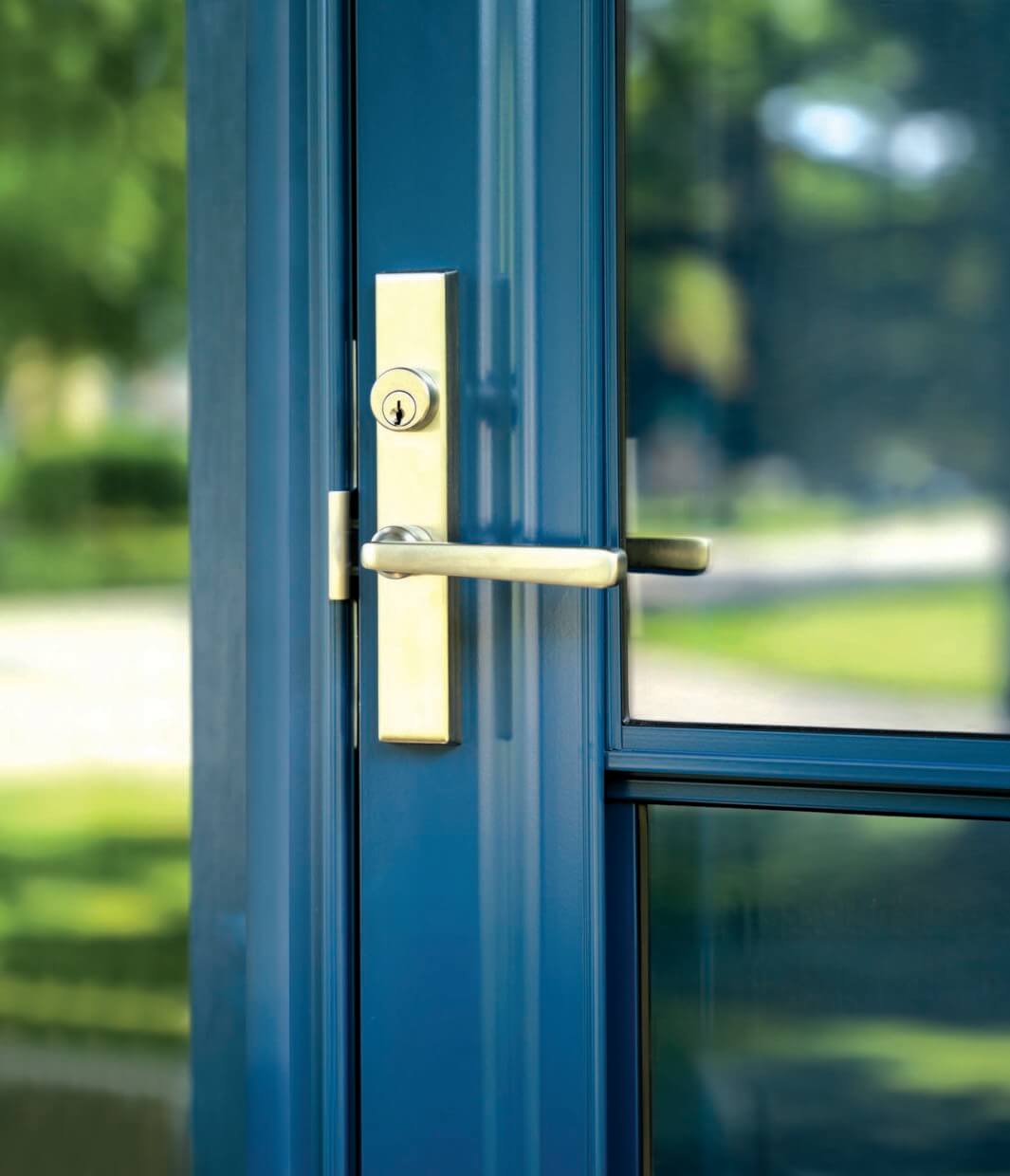 Close-up of a blue-painted exterior door with a brushed brass handle and lockset, set against a glass panel reflecting greenery outside.