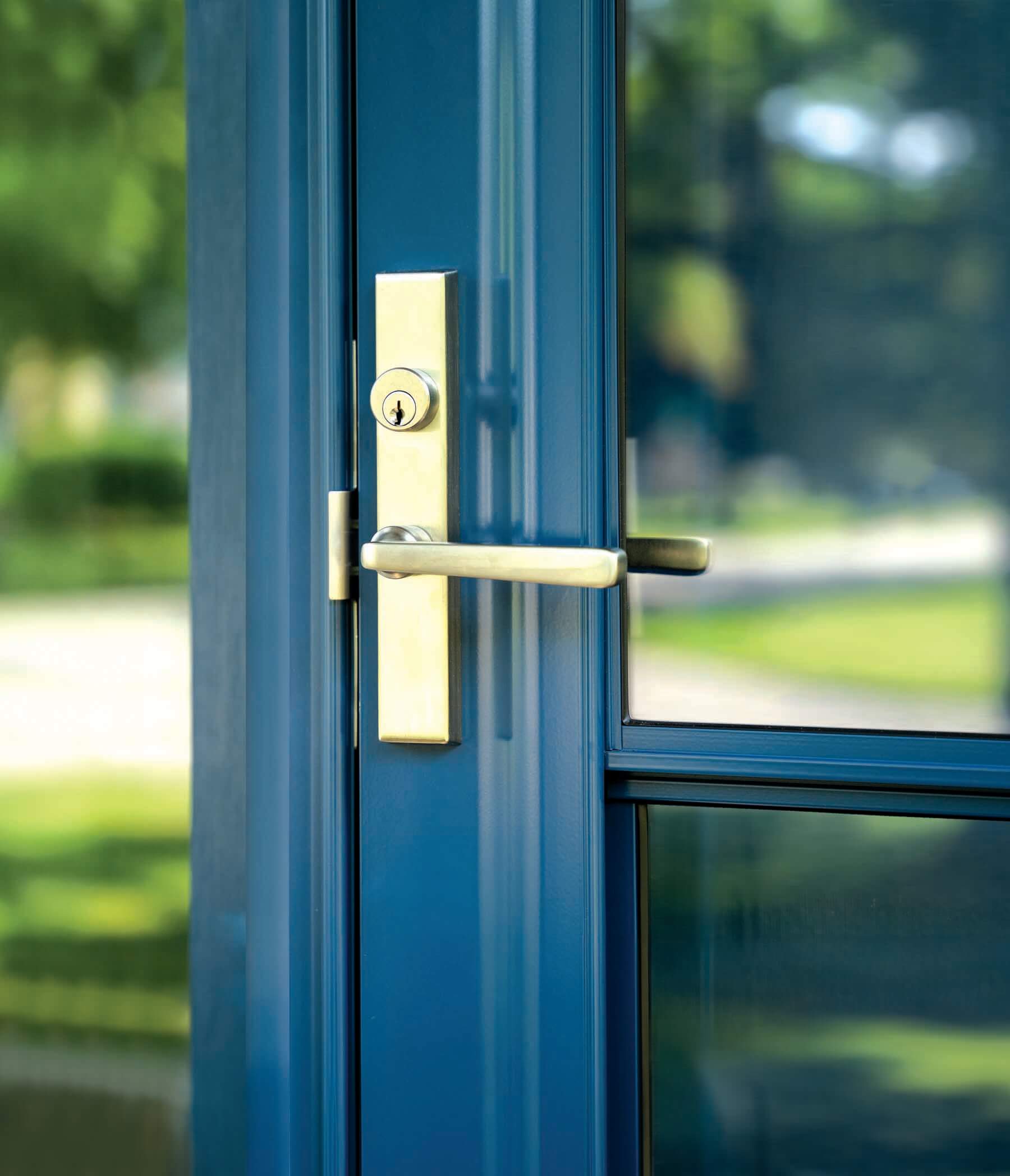 Close-up of a blue-painted exterior door with a brushed brass handle and lockset, set against a glass panel reflecting greenery outside.