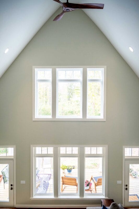 Bright vaulted living space with a high ceiling and wood-bladed ceiling fan, featuring a stacked arrangement of large white-trim windows that flood the room with natural light and overlook a deck with outdoor seating and surrounding trees.