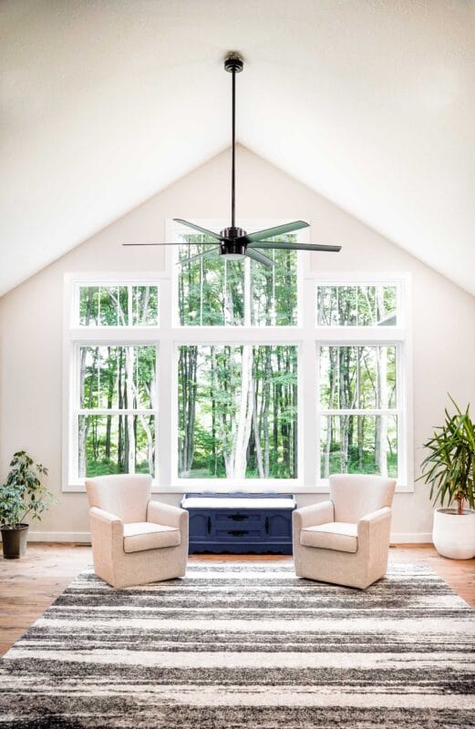 Bright living room with a vaulted ceiling featuring a black ceiling fan, large grid-style windows overlooking a wooded yard, two light upholstered armchairs facing a blue storage bench, and a patterned area rug on hardwood floors.