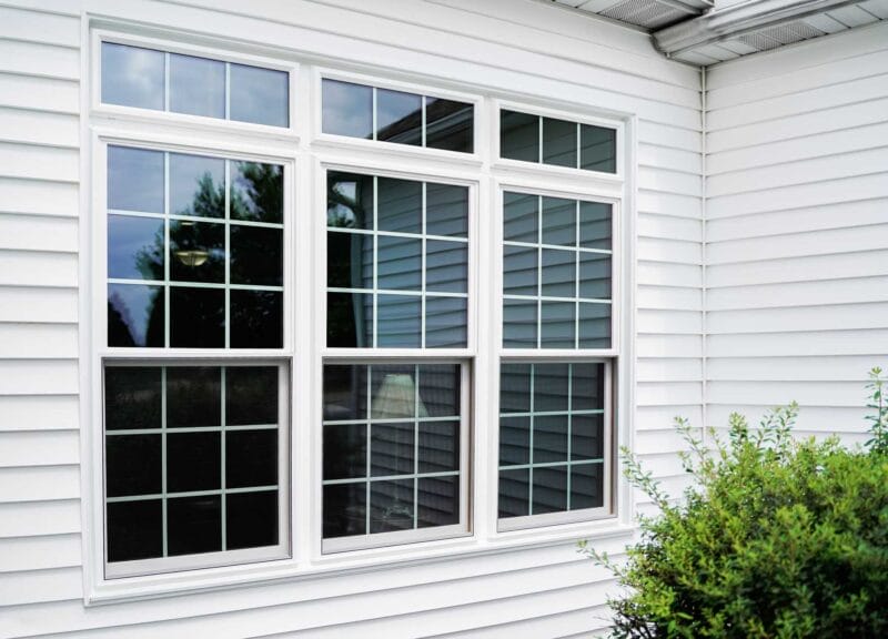Four-pane exterior window with white trim and grid-style divided glass, set into white vinyl siding with a small shrub below the sill.