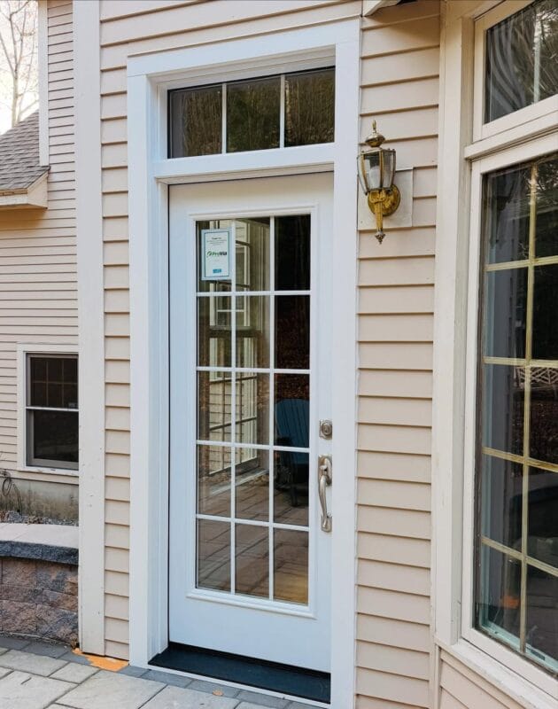 White French-style exterior door with divided glass panels and transom window, installed in beige siding with white trim and an adjacent wall-mounted lantern.