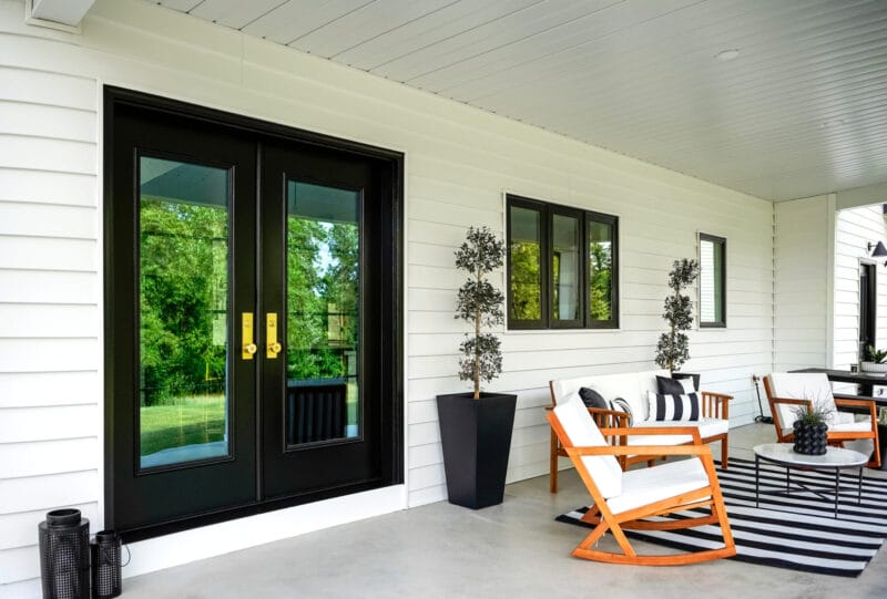 Black double entry doors with glass panels and brass hardware on a covered porch with modern outdoor seating and striped rug.