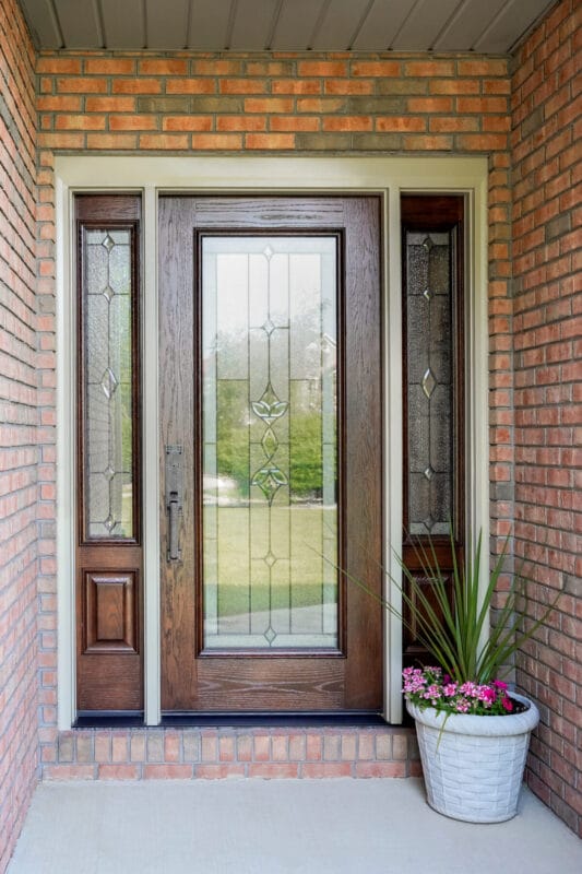 Wood front entry door with decorative glass panel and sidelights, set in a brick exterior with a potted plant beside the entrance.
