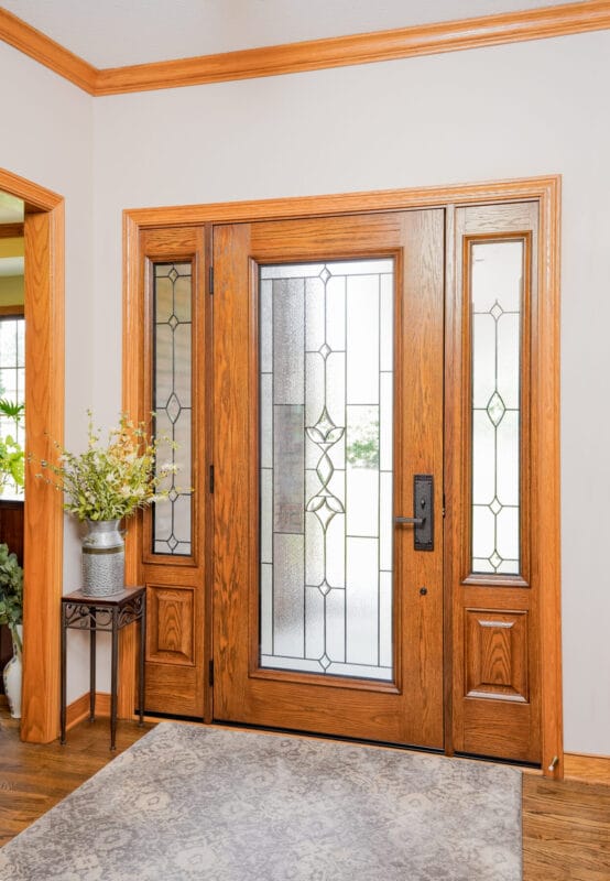 Interior view of a wood front door with decorative glass and sidelights, surrounded by matching trim and hardwood flooring.