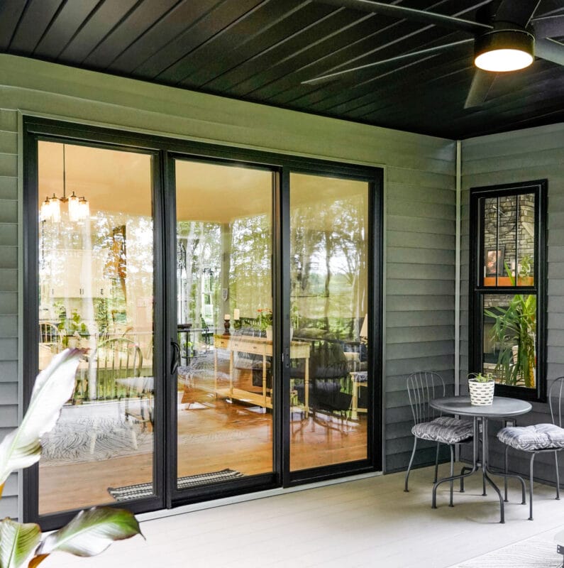 Covered patio with black-framed sliding glass doors, showing a view into a bright dining area, with a small table, chairs, and potted plants nearby.