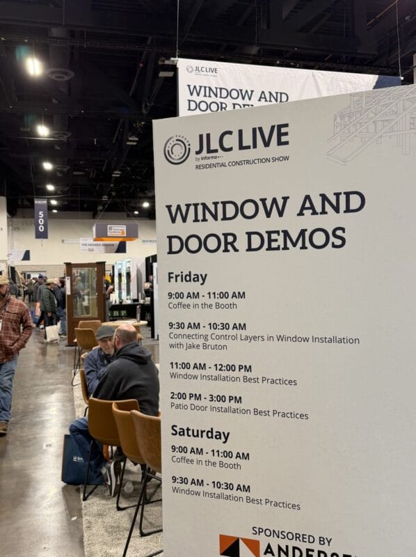 Large event sign at a construction trade show displaying schedule for window and door installation demos with attendees in the background
