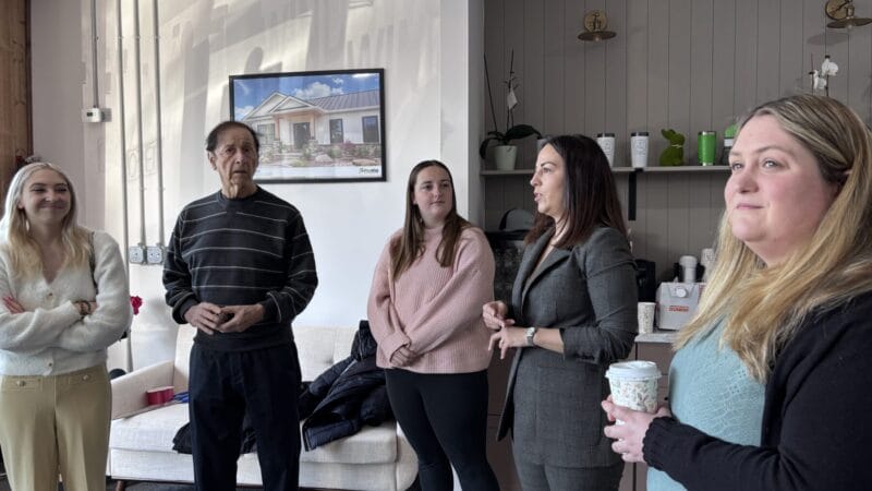 Group of people gathered inside a showroom, listening as a woman speaks, with coffee cups, branded merchandise, and a window display visible in the background.
