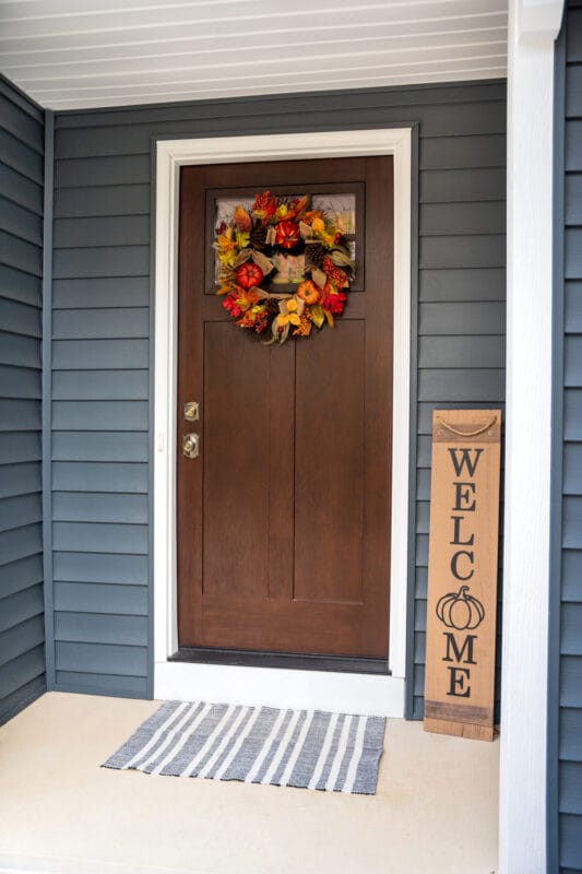 Front entry with a stained wood door, fall wreath, blue siding, and a vertical “welcome” sign beside the doorway.