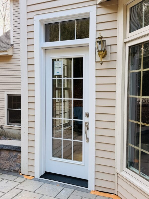White exterior door with divided glass panels and a transom window above, set in beige siding with white trim and a wall-mounted lantern beside it.