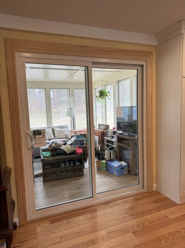Interior view of a sliding glass door with natural wood trim, leading into a sunroom filled with furniture, a TV, and large surrounding windows.