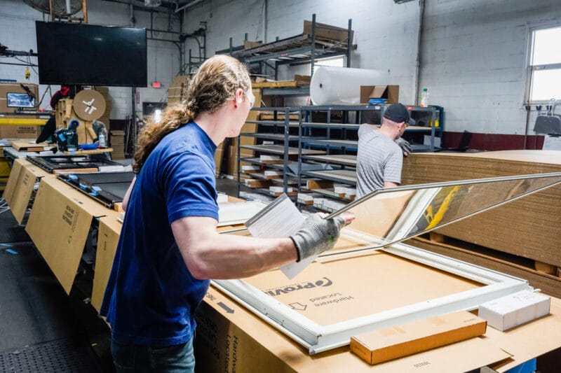 Worker reviewing and assembling a window frame on a workbench while checking paperwork in a manufacturing facility