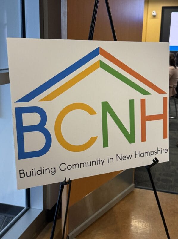 BCNH sign on an easel displaying “Building Community in New Hampshire” with a colorful house-shaped logo at an indoor event venue.