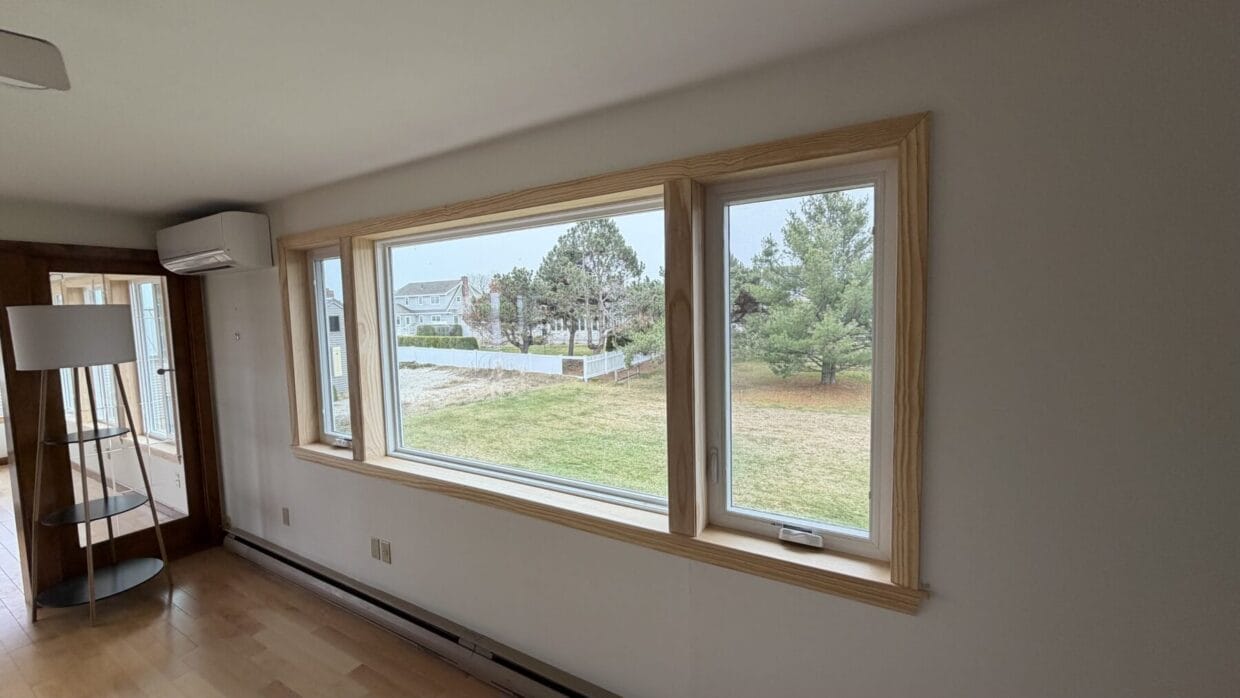 Large picture window with natural wood interior trim installed in living area overlooking grassy backyard and coastal neighborhood homes