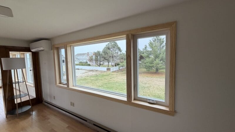 Large picture window with natural wood interior trim installed in living area overlooking grassy backyard and coastal neighborhood homes